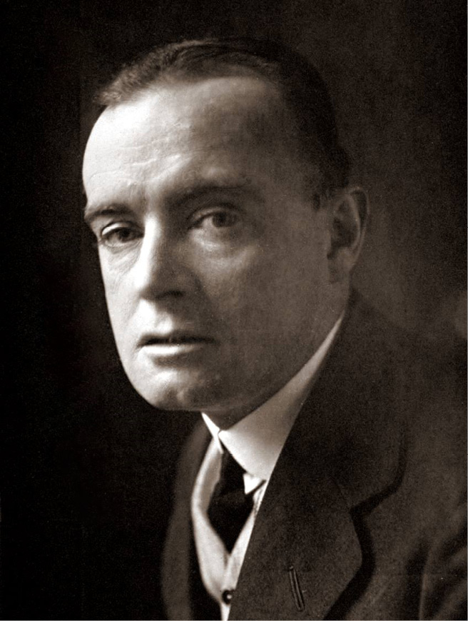 A black and white photograph of a man, 'Saki', dressed in a black suit, white shirt and black tie. He is looking towards the camera and his face is half in shadow.