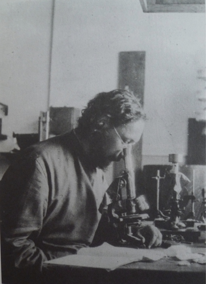 A photograph of a man with short, dark, curly hair and wire-rimmed glasses looking down a microscope.