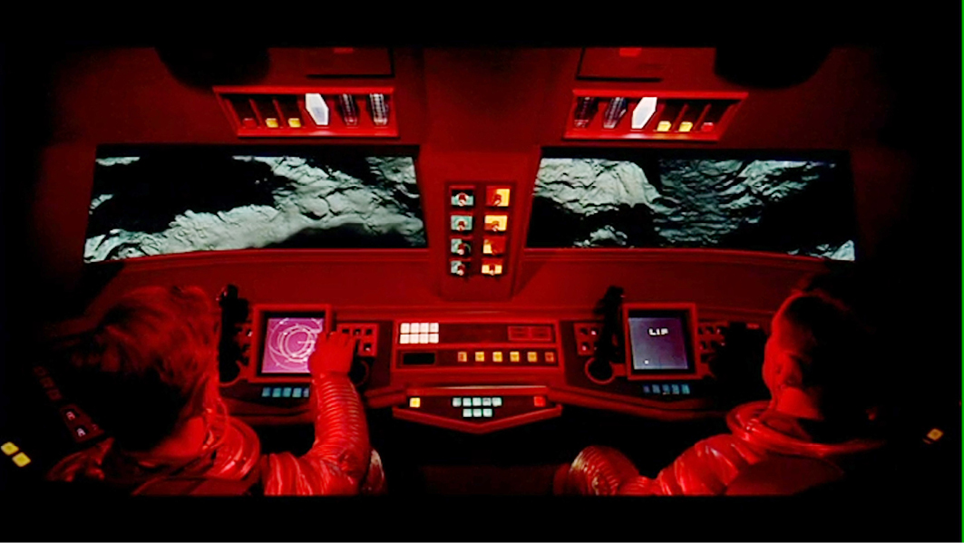 A cockpit view from the movie “”2001: A Space Odyssey”” showing two astronauts operating controls in a red-lit space shuttle, navigating over a planetary surface.”,