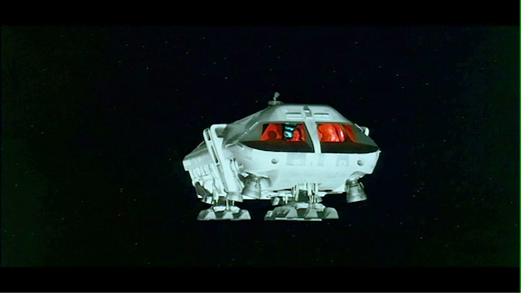 A scene from “”2001: A Space Odyssey”” showing an exterior view of a white space shuttle floating in the dark vacuum of space.”,