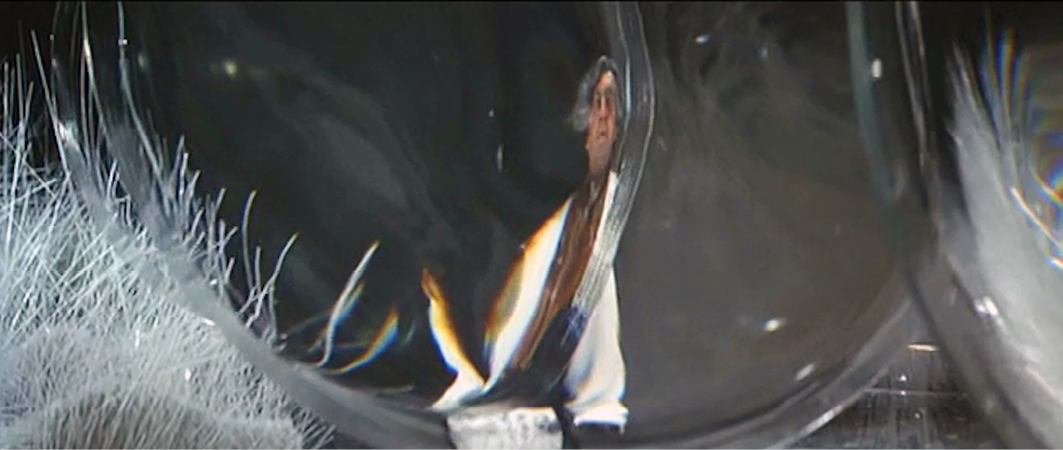 A distorted view of a character seen through a warped glass-like structure in the film “”Barbarella”” (1968). The character appears reflected and bent, creating an abstract, almost psychedelic visual effect.”,