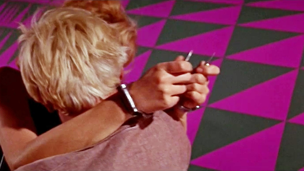 A close-up shot from ‘Modesty Blaise’ where two characters are embracing. The woman’s hands hold a small sharp object behind the man’s back, creating a sense of tension. The purple and black patterned wall serves as the background.”,