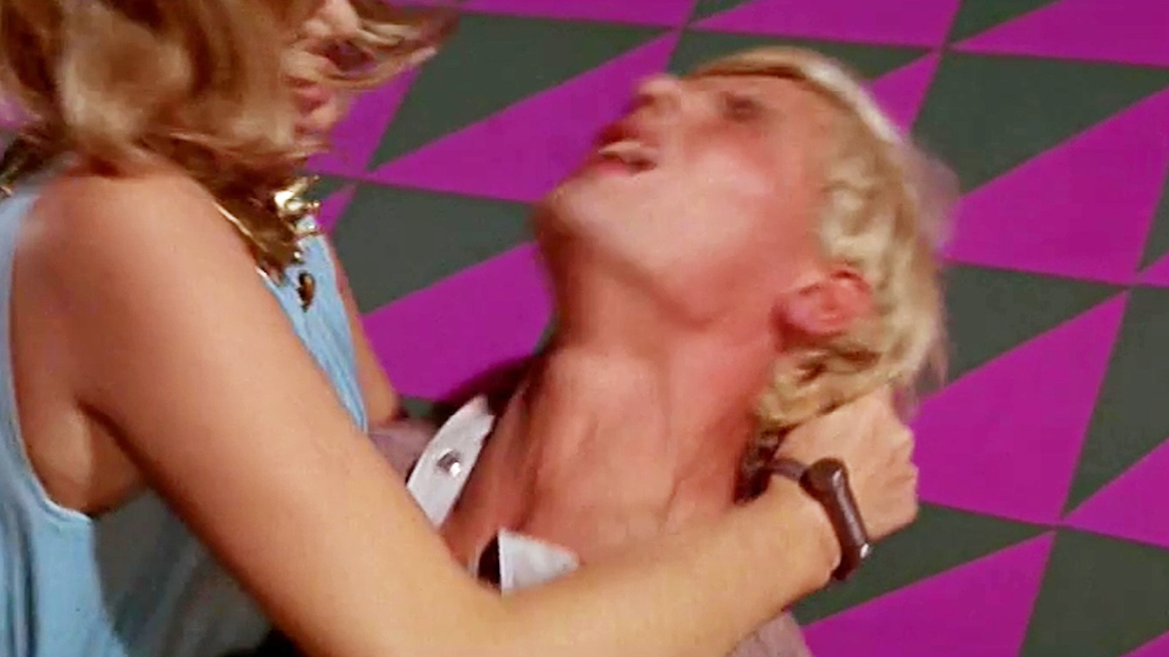 A dramatic shot from ‘Modesty Blaise’ featuring Modesty grabbing a man by the neck, her expression intense. The purple and black triangle-patterned wall is prominent in the background, adding to the dynamic tension of the scene.”,