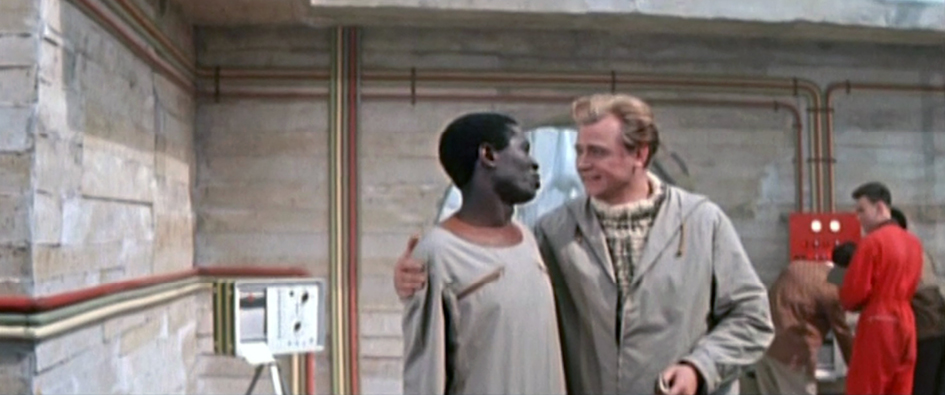 A scene from the 1960 East German-Polish science fiction film ‘The Silent Star’ (Der Schweigende Stern). The image shows two men embracing in a futuristic setting, with a greyish industrial background. This film is a portrayal of an international team of astronauts on a mission to Venus.”,