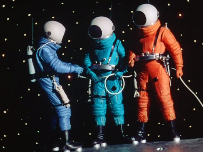 A scene from the 1950 film ‘Destination Moon,’ showcasing three astronauts in space suits of blue, teal, and orange floating in space. The background is a starry sky, and the astronauts are connected by cables.”,