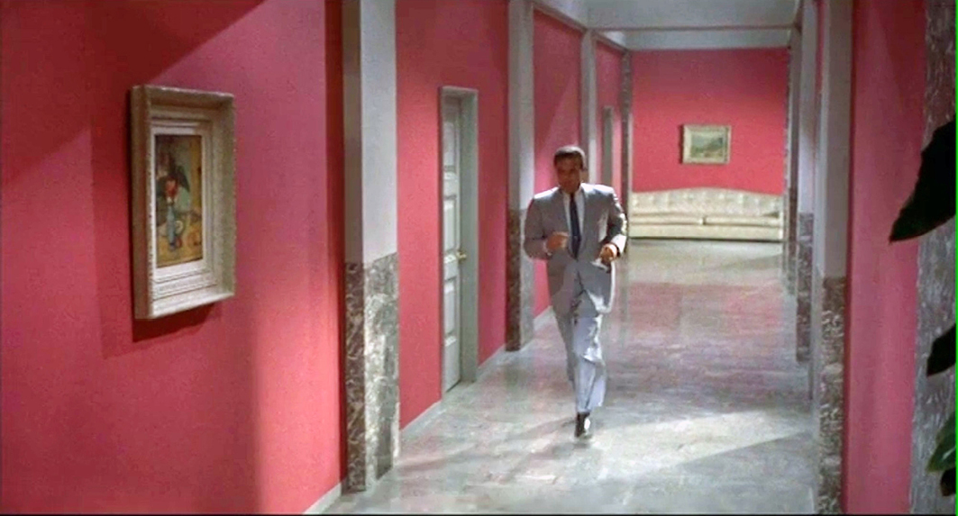 A hallway scene from the 1956 film “”Written on the Wind,”” directed by Douglas Sirk. A man in a gray suit walks down a brightly lit corridor with red walls, gray doors, and artwork on the walls. The polished floor reflects the stylish setting.”,