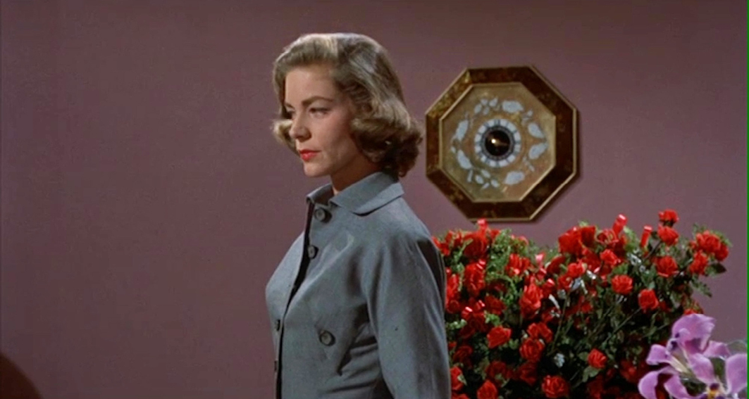 A scene from “”Written on the Wind”” showing a woman in a light grey jacket standing near a decorative mirror and a vibrant arrangement of red roses. The woman appears poised and deep in thought.”,