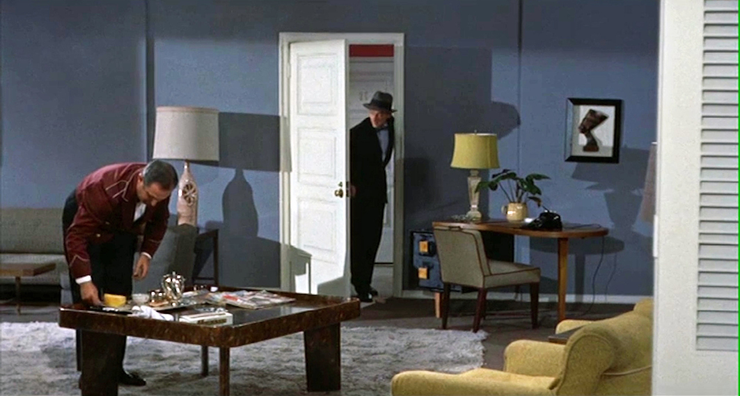 A hotel room scene from “”Written on the Wind”” where a man dressed in a suit and fedora enters the room while the hotel staff tidies up a table. The room has blue walls, mid-century modern furniture, and a comfortable atmosphere.”,