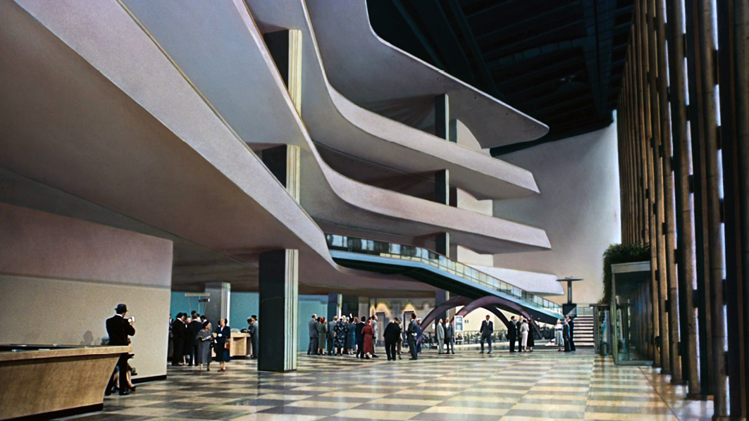 A wide shot of the iconic modernist interior from “”North by Northwest”” showing a futuristic lobby with sweeping architectural lines and people gathered in the background. The design emphasizes sleek, curved forms and open space.”,