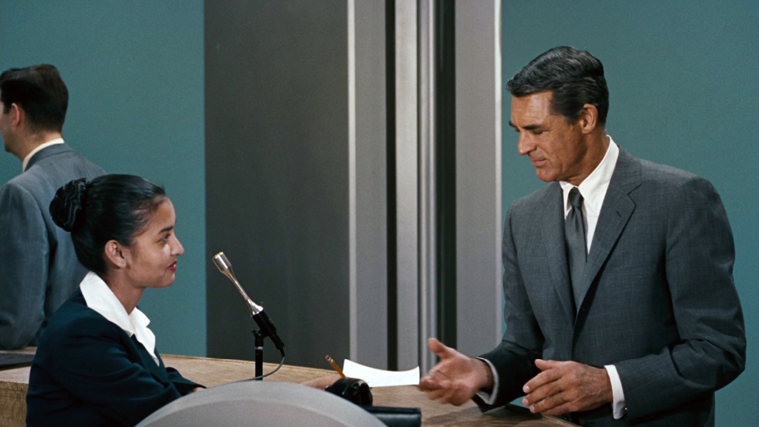 A close-up from “”North by Northwest”” featuring the protagonist, played by Cary Grant, interacting with an airport clerk at the ticket counter. The protagonist is dressed in a grey suit, appearing calm yet concerned.”,