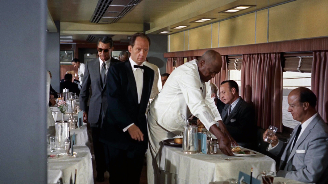 A scene from the dining car in “”North by Northwest”” where the protagonist, disguised with sunglasses, walks through a formal dining area filled with passengers and waitstaff. The car is lively, with white tablecloths and suited diners.”,