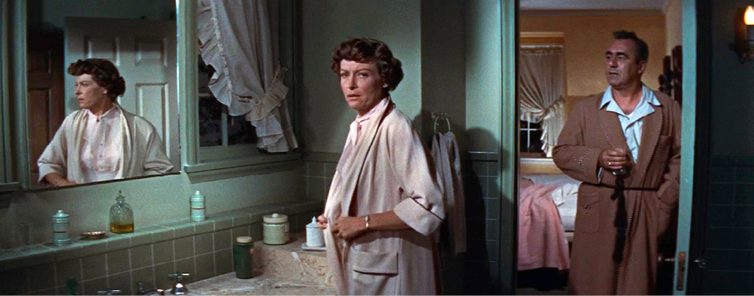 A scene from “”Rebel Without a Cause”” featuring two characters standing in a bathroom. The woman, wearing a light pink robe, looks concerned as she faces her reflection in the mirror. A man, wearing a brown robe and holding a drink, stands in the doorway of the bedroom behind her.”,