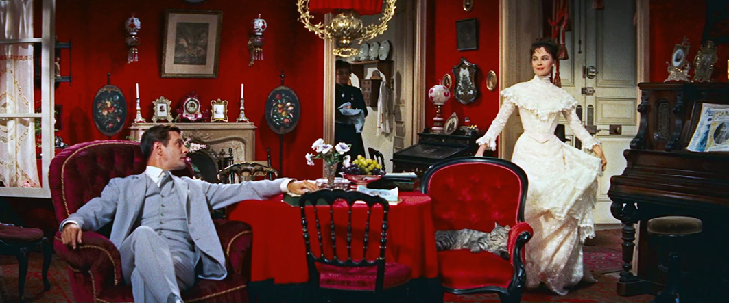 A man in a light grey suit sits on a red velvet armchair, while a woman in an elegant white dress walks into the red-decorated Victorian parlor. The room is filled with luxurious decor and old-fashioned furniture. This scene is from the musical film ‘Gigi’ (1958).”,