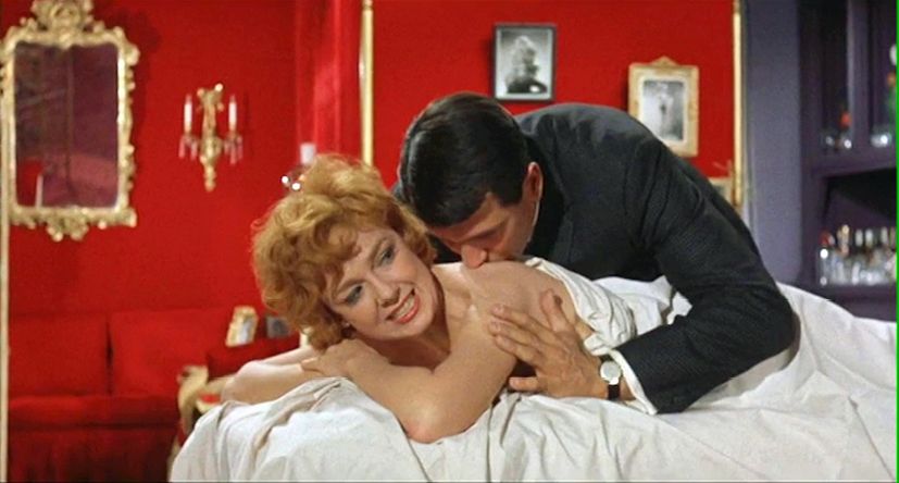 A man kisses a woman on the shoulder while she lies on a bed smiling. The room is decorated in deep red with gold accents. This scene comes from the romantic comedy ‘Lover Come Back’ (1961), showcasing an intimate moment.”,