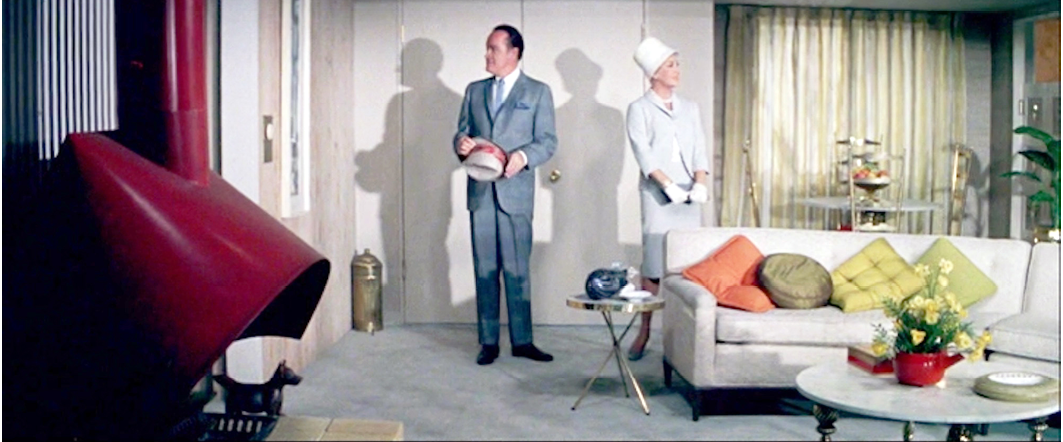 A man and woman stand in a mid-century modern living room, the man holds a hat while observing the room. The living space is minimalistic with muted tones and colorful pillows. This scene is from the 1961 film ‘Bachelor in Paradise’.”,