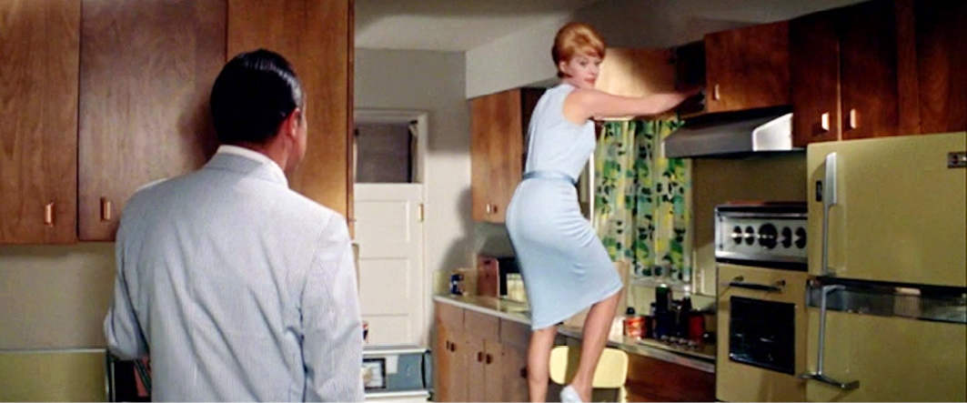 A man in a light-colored suit observes a woman standing on a chair, reaching for something on the kitchen cabinets. The kitchen has wood-paneled cupboards and a yellow refrigerator. This is a scene from ‘Bachelor in Paradise’.”,