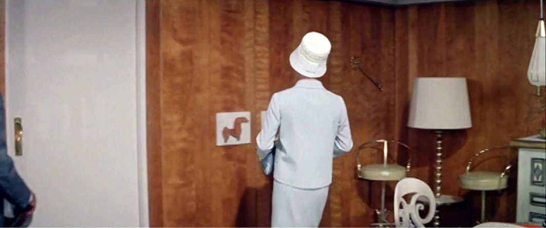 A woman in a white hat and suit looks towards a wood-paneled wall, her back to the camera, in a mid-century modern room. The wall has a small horse artwork and modern furniture. This scene is from ‘Bachelor in Paradise’.”,