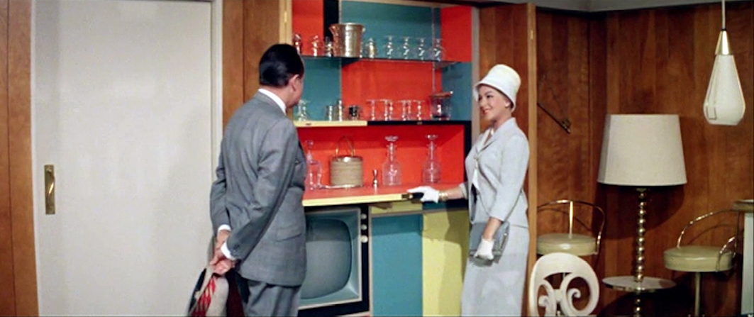 The same woman in a white hat and suit smiles at a man while showing him a colorful, hidden bar space in the wood-paneled room. Glassware and a television are visible. This is from ‘Bachelor in Paradise’.”,