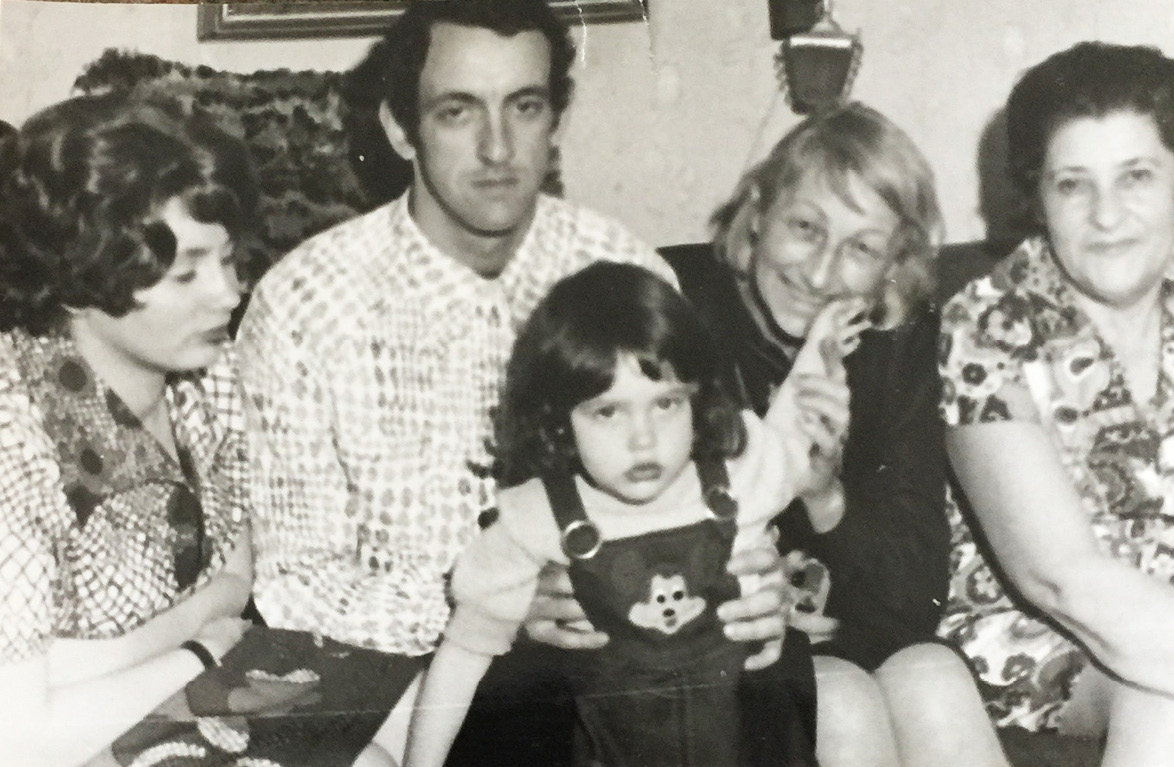 A black and white photograph from 1977 with a girl with dark hair of about 3 years old, wearing overalls with a mouse on it. She has a pensive look on her face and one of her hands is raised. Behind her are four adults seated on a couch. The one on the left is her mother who is looking at her. Next to the mother is the girl’s father, grandmother, and one more person.