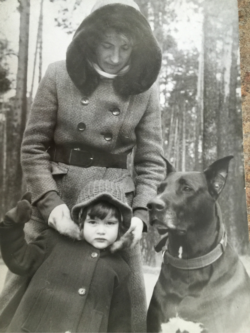 A black and white photo from 1977 of a girl of about 3 years old, wearing a hat and a dark button-up coat. Behind her is a middle-aged woman wearing a coat with a furry hood. At their side is a large black great dane with some white on his chest. The dog is seated with a collor on his neck.