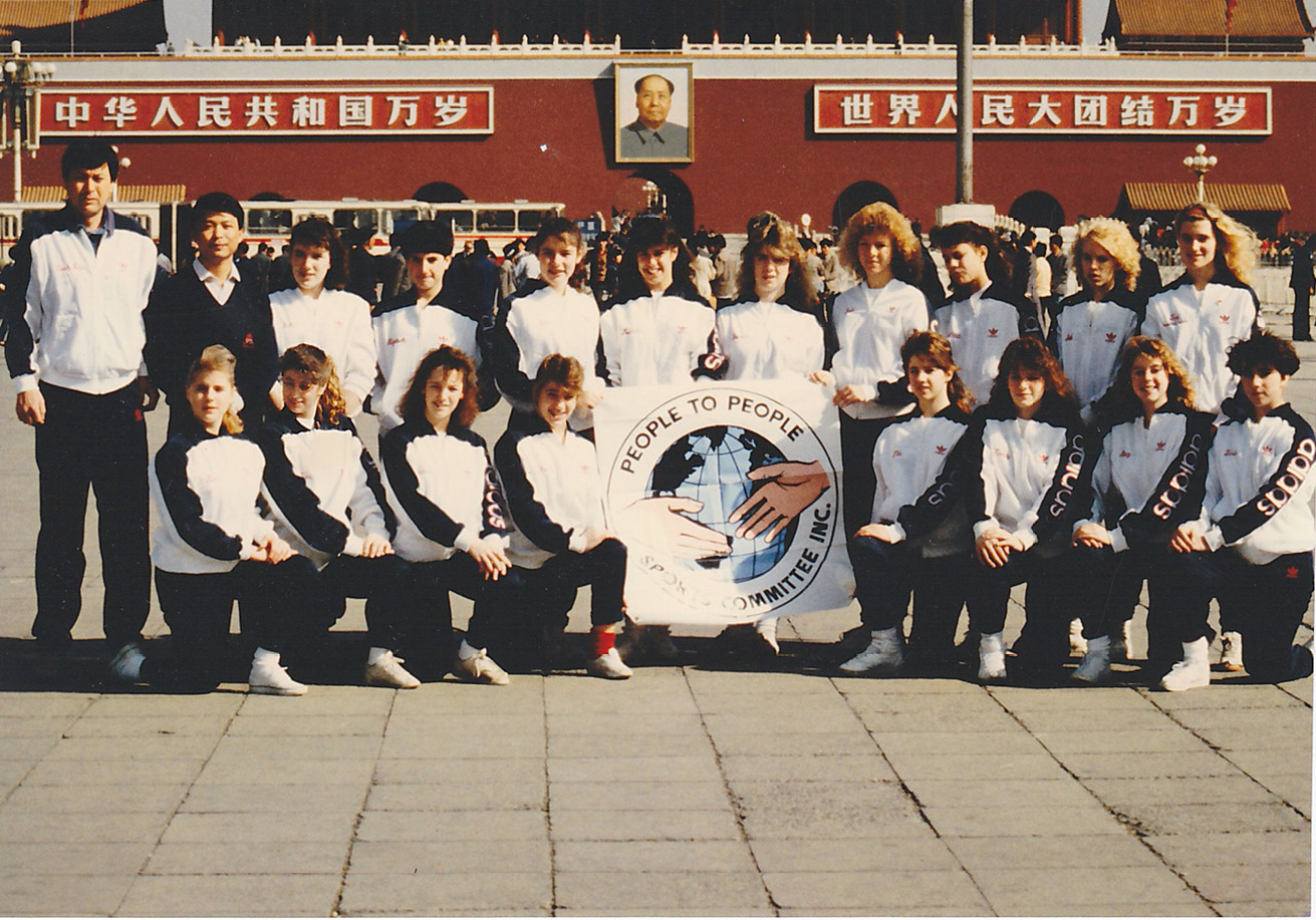 A group picure with nineteen peope, mosly highschool girls, as well as their gymnastics coach and tourguide. There are eight girls kneeling in the front and nine girls standing behind them along with the two men. The girls in the middle are holding a sign that says “People to People Sports Committee Inc.”. Inside the sign, on the background, there is a map of the world; in its foreground, there are two hands reaching toward one another for a handshake. The team is standing in Tianamen Square in Beijing, China and the girls and their coach are all wearing dark pants and a Addidas shirt that has dark sleeves and a white front.