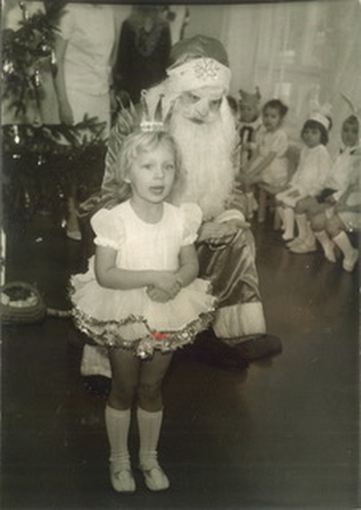 A photograph from a New Year’s party at the kindergarten. A girl of about 4 or 5 years old is standing in front of the Father Frost (Santa Claus). The girl has short blonde hair. She is wearing a white snowflake costume handmade by her grandmother and parents. The skirt is made from multiple layers of starched white gauze with silver garland attached to the edges of each layer. The crown is made from silver lining of a cigarette box. The girl’s mouth is open as if she is talking or reciting a poem. The Father Frost listens carefully. In the background, other children are watching the girl.
