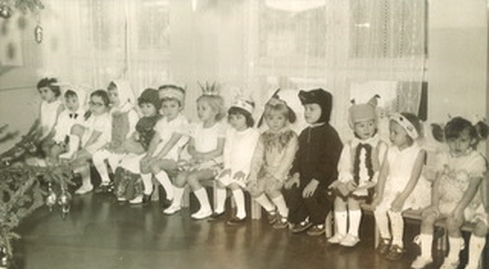 A photograph from a New Year’s party at the kindergarten. The children of about 4 or 5 years old are sitting on a bench. They are wearing different costumes, incuding a bear, hare, fox, Little Red Riding Hood, snowflake, and others. Some girls are wearing white bows or crowns. The girl in a snowflake costume is sitting with her hands crossed and resting on her knees. She is seventh from the left.