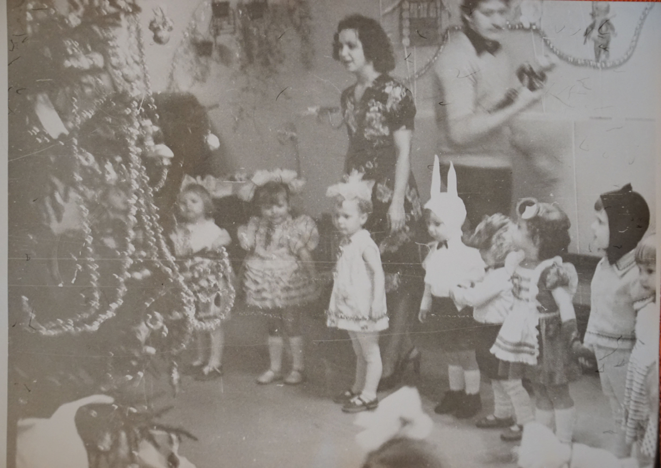A photograph from a New Year’s party at the kindergarten. The children are dancing and singing around a decorated Christmas tree. The boys are mostly wearing bunny costumes and the girls are dressed as snowflakes. Only one girl is wearing a frog costume. She is holding hands with other children and her gaze is focused on the Christmas tree. The kindergarten children is standing behind the children with her mouth open, most probably she is singing with children. A photographer is passing behind her.