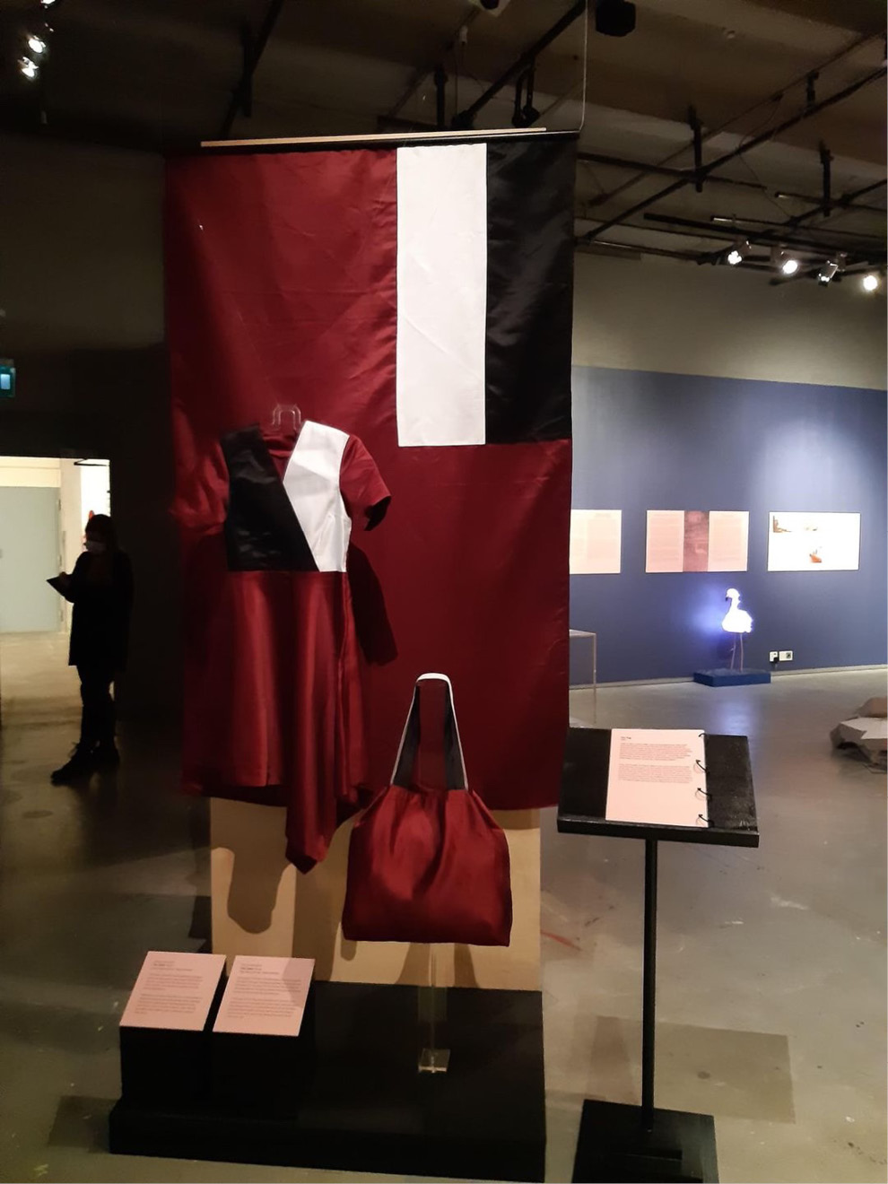 A color photograph from a museum exhibit, displaying a hand-made dress and a grocery bag made out of a Georgian flag. The dress, which is dark red with white and black elements, is displayed on a mannequin. The bag, which is dark red with black-and-white handles, is placed to the right of the dress. In the background hangs a large Georgian flag, which is dark red with a canton horizontally divided in black-white.
