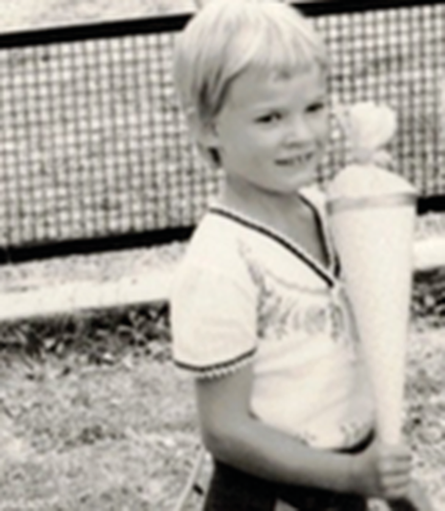 A black-and-white photograph of a six-year old girl with short blonde hair, wearing a white blouse with emboirderted decorations. The girl is smiling and hodling what appears to be a present in a form of a large white cone.