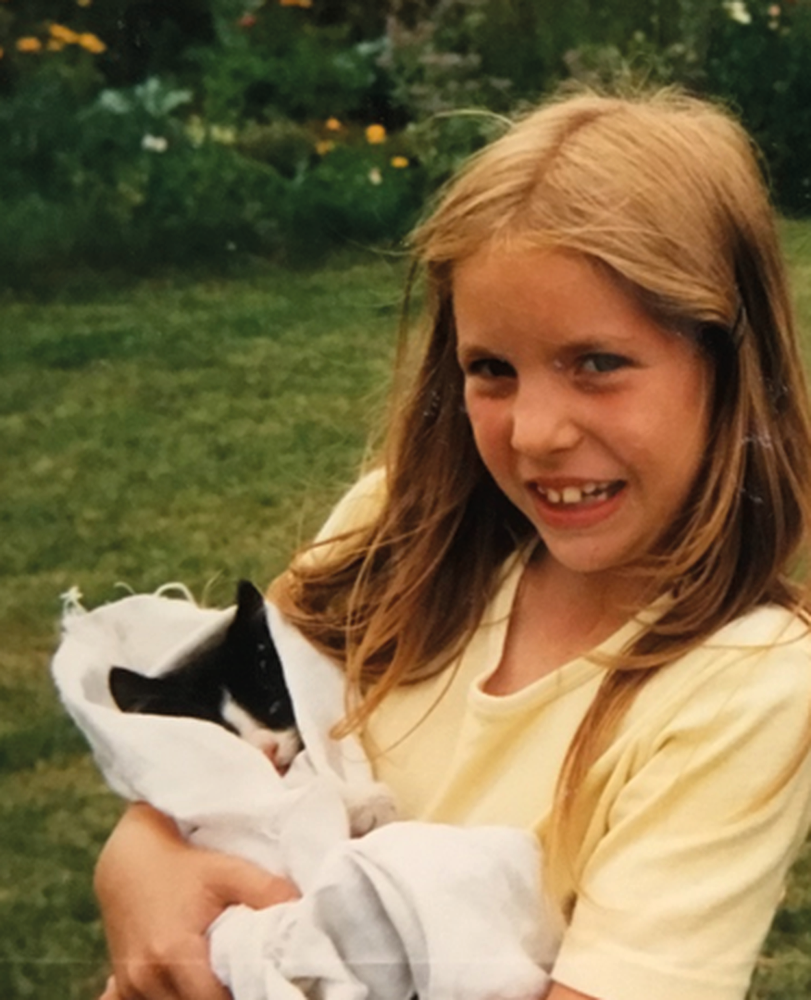 A color photograph of a young girl, approximately eight or nine years old, with long straight hair. The girl is wearing a yellow t-shirt. She is smiling and holding a small black and white kitten, which is wrapped in white cloth.