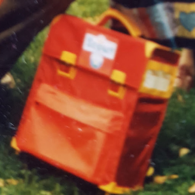 A color photograph of a red school backpack with yellow buckles, handle, and edges. The backpack is rectangular, with distinctly sharp edges.