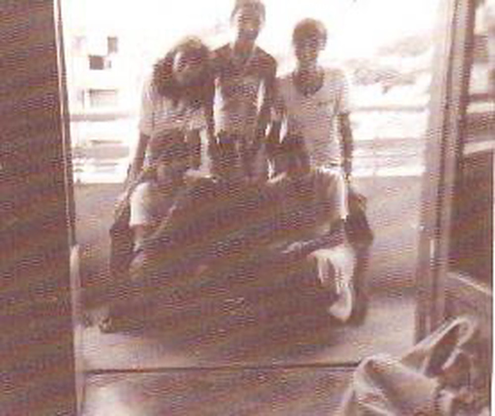 A photograph in sepia of six young students, mostly girls on a balcony. The picture was taken from inside the apartment and the image is overexposed and blurry. The faces are hardly distinguished, but some seem to be smiling. Three persons are standing and three girls are bending on their knees. The view shows some buildings in the background.