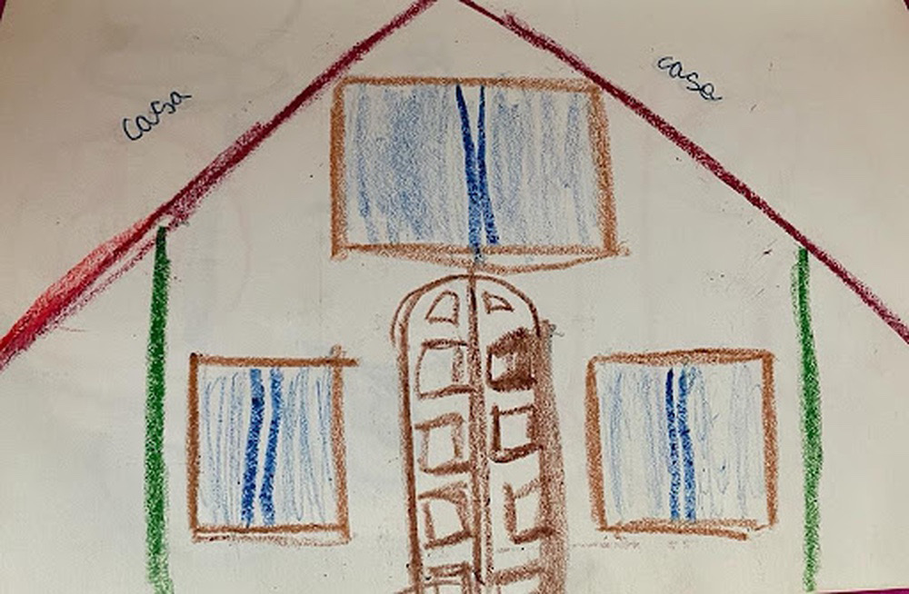 Drawing of a gabled-roof house with a front door, two windows on the ground floor and a larger window on the first floor. The lines are simple and were made with crayon colors. The roof lines are red, the walls are green, the door is brown and shows several wood panels, and the windows are drawn in blue, with curtains closed. Above the gabled roof there are the words “casa” (house/home) on both sides.