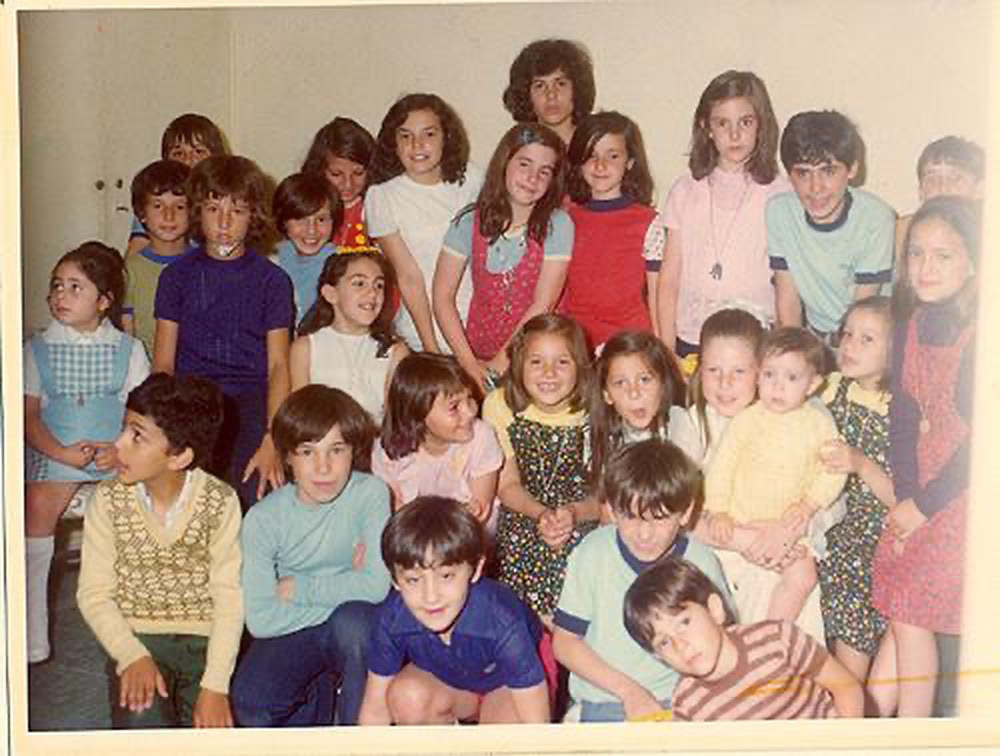 Color photograph showing a large group of children of different ages. One girl on the second row has a party hat. Most children are smiling at the camera. On the left side, a boy and a girl are looking at something that is happening outside the frame. The author is on the upper row, with a shy smile, red jumper and light blue T-shirt. Her sister is right above her; only her head is visible, and she looks serious.