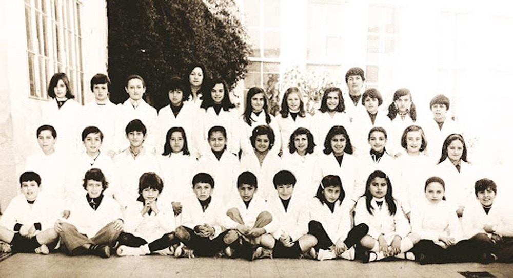 Black and white school photograph of a group of students with two teachers standing behind the upper row of students. The 32 students are shown in three rows, the first one seating with their legs crossed, the second on their knees, and the third one standing. All the students are wearing a white apron, the typical school uniform in Argentina. The picture was taken on a schoolyard; the tiled floor is visible, as some plants and classroom windows on the background and on the left side. Most students are smiling and their look seems relaxed. The right side of the photograph seems overexposed with light, but all bodies are visible.