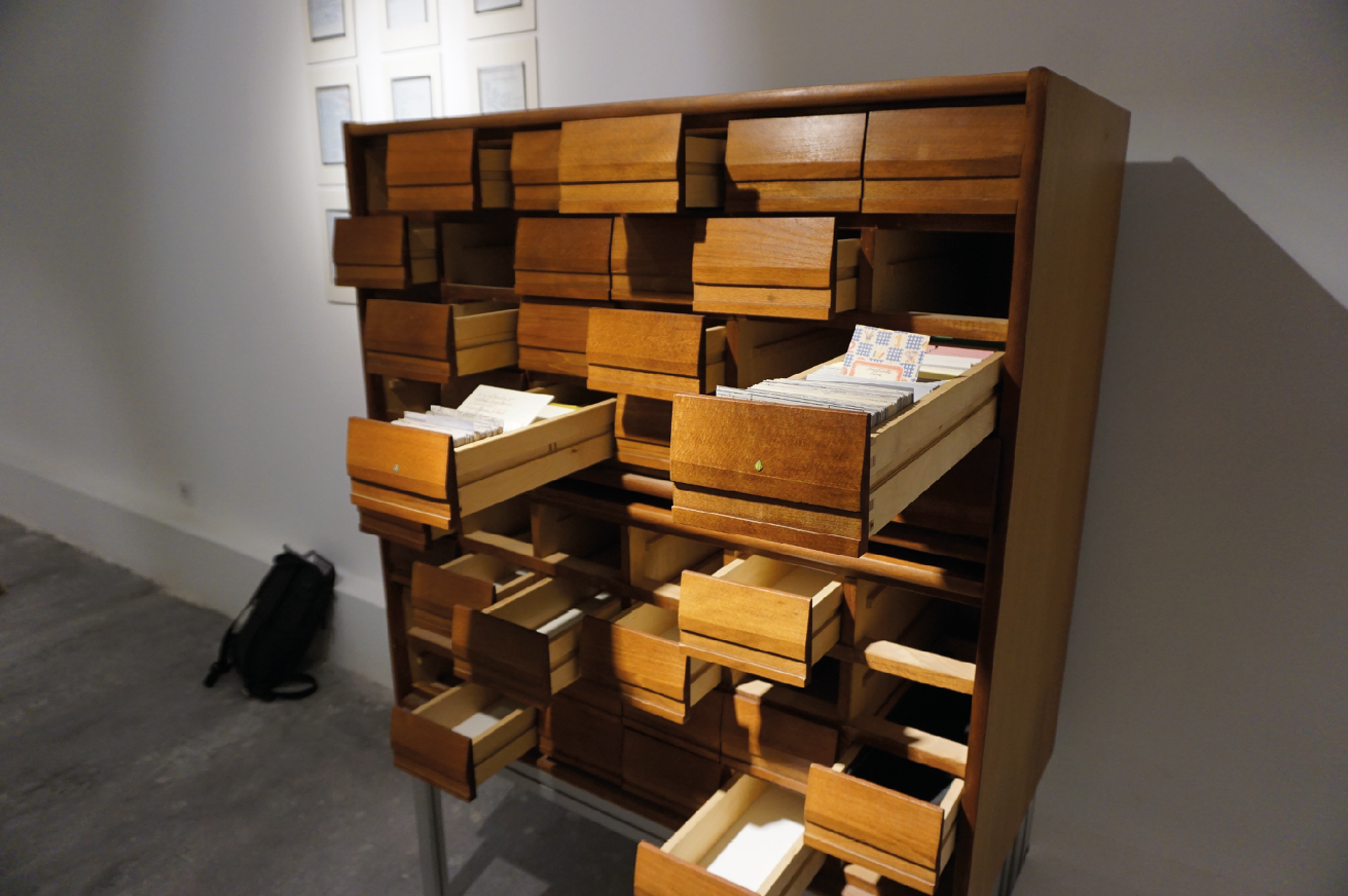 In the exhibition room, an oak old-style library catalogue cabinet stands by the wall. Half of the drawers are pulled out to different extents from the cabinet and some draweers are missing. Some of the drawers are empty and some are filled with library catalogue entries. On the right hand of the cabinet, in the open drawer, a 1990s style school notebook is turned vertically so it is visible among the other orderly library entries.