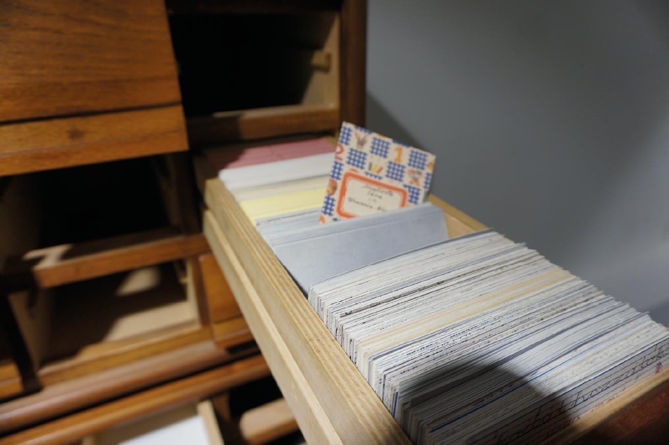 An old oak library catalogue is pulled out from the cabinet and is filled with paper catalogue entries. In the middle of the drawer, among the catalogue entries, a 1990s style school notebook is turned vertically so that its face is visible. The schoolbook has a first year schoolbook style of decoration with a label on it with the name of the student and the class.