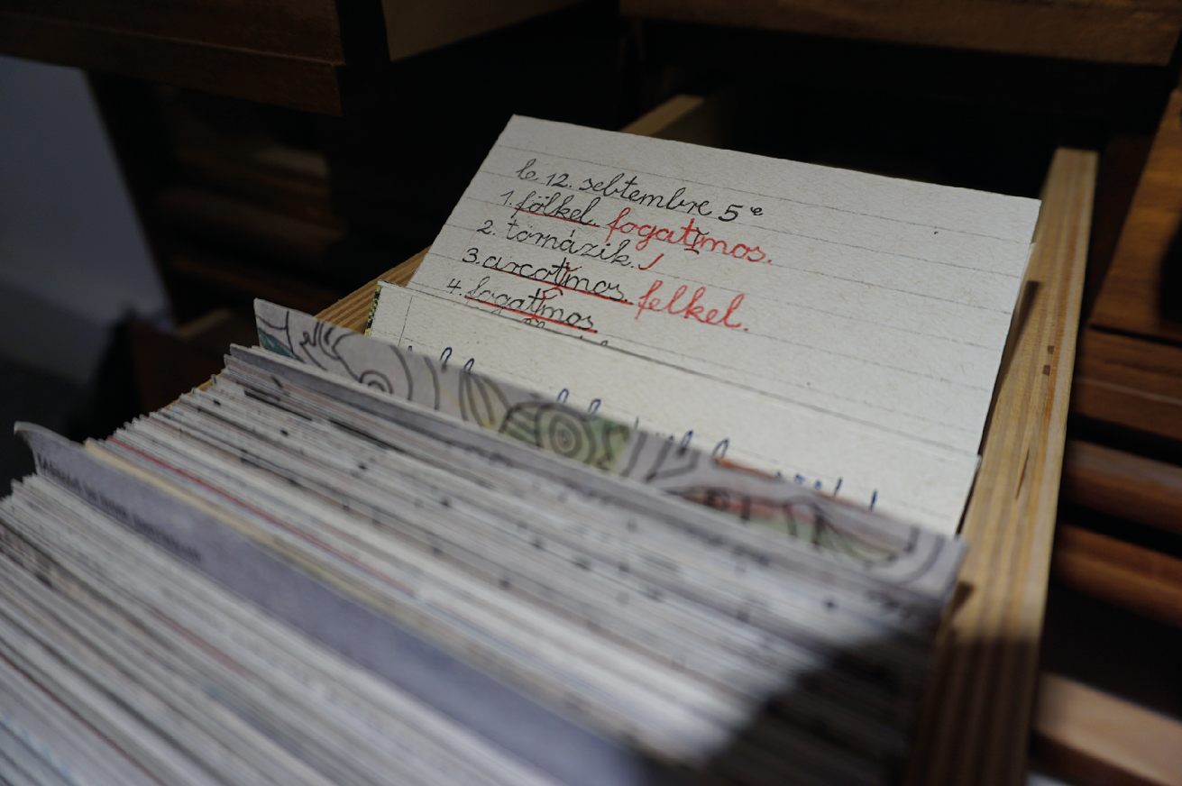 A drawer is pulled out from the woden library catalogue cabinet pictured up close. The drawer is full and one entry is pulled out at the back which has five lines of childlike writing. The writing is a list of activities composing a morning routine. Two lines of the routine are underlined in red pen and and other words are written in red pen next to those. The new words in red pen are a playful mixing up of the routine.