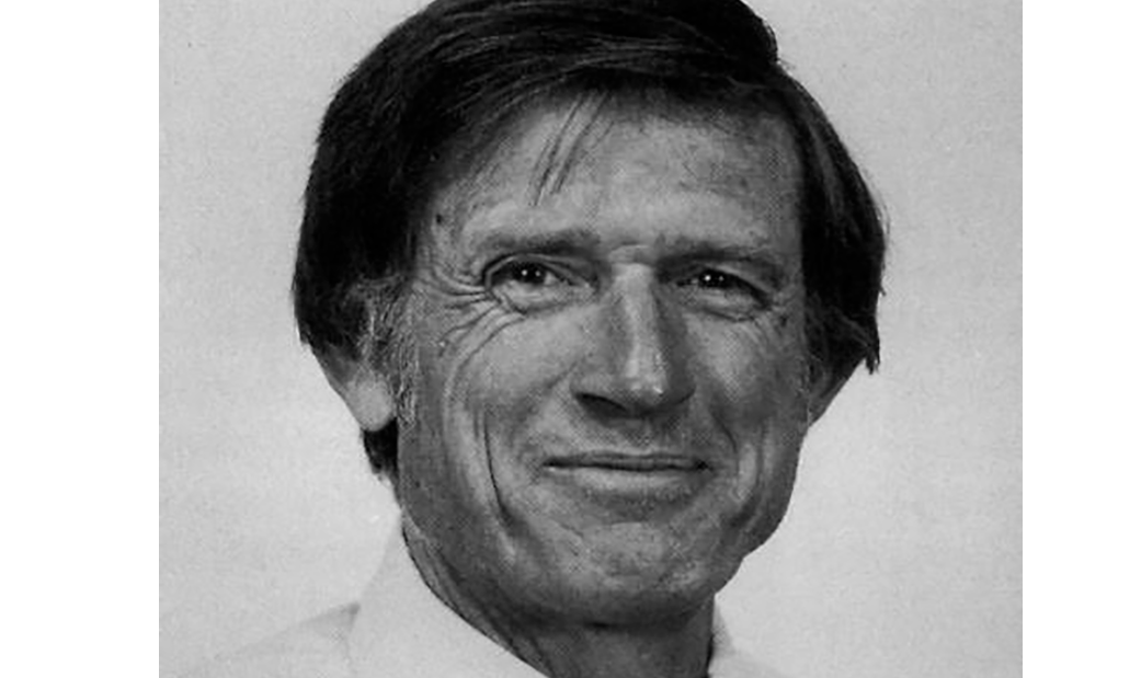 A black-and-white headshot of a light-skinned man with short, dark hair, slight sideburns and dark eyes. He smiles at the camera, face crinkling around his eyes, and wears a white, collared shirt.