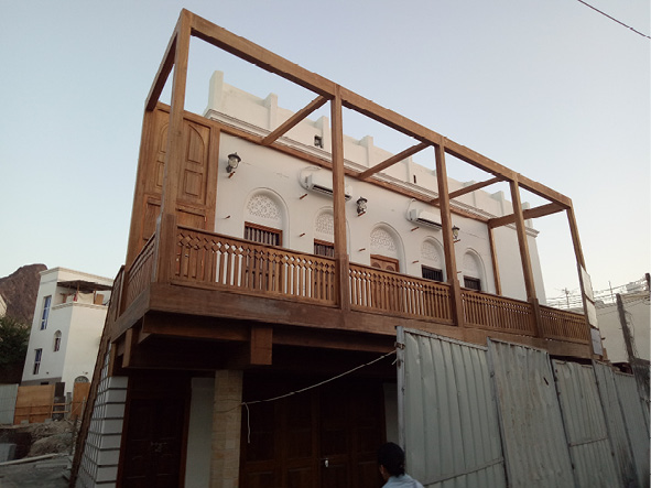 Image of a building under development featuring a white facade with a wooden balcony, a large wooden gate, and multiple doors