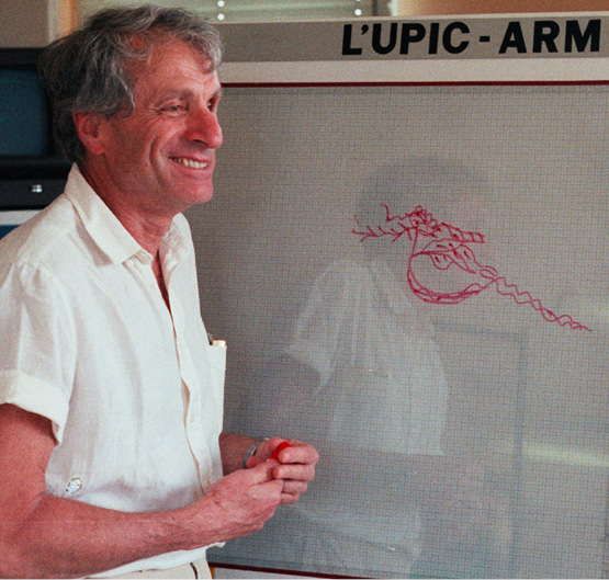Iannis Xenakis wearing a light-colored shirt stands next to an electronic drawing board labeled “L’UPIC-ARM.” He is holding a red marker and smiling, with a sketch of abstract shapes on the board. A computer monitor is visible in the background.