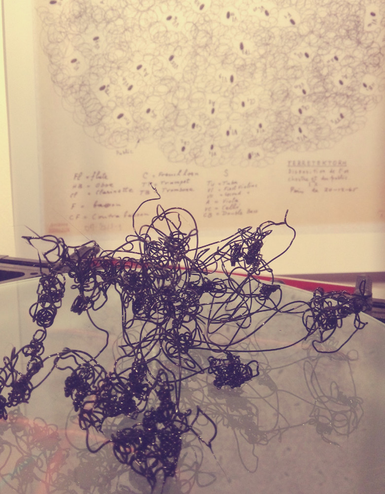 A complex and tangled black wire sculpture is displayed on a glass surface, with its reflection visible below. In the background, a large piece of paper contains intricate abstract line drawings and handwritten notes describing Xenakis’s work Terretektorh.