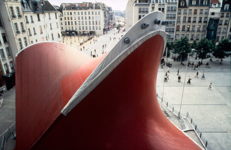 Aerial view of a large, hyperbolic-paraboloid construction in red fabric placed in a city square.