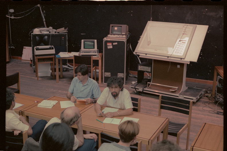 Two men sitting side by side at a table in a classroom facing a group of people whose heads can be seen in the bottom of the picture. Behind the men is a large drawing board on a plinth, two old-fashioned, green-screen computer terminals, and a printer on a table in front of a large JBL loudspeaker.