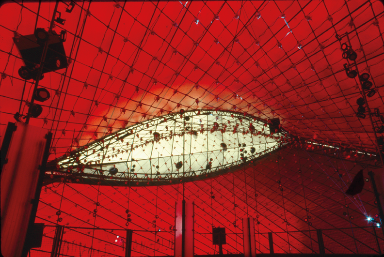 The inside of a large tent-like structure in red fabric, with an eye-shaped portion in white fabric at the far end. The fabric is criss-crossed with cables supplying an array of small lights. A number of loudspeakers on stands are visible around the space.