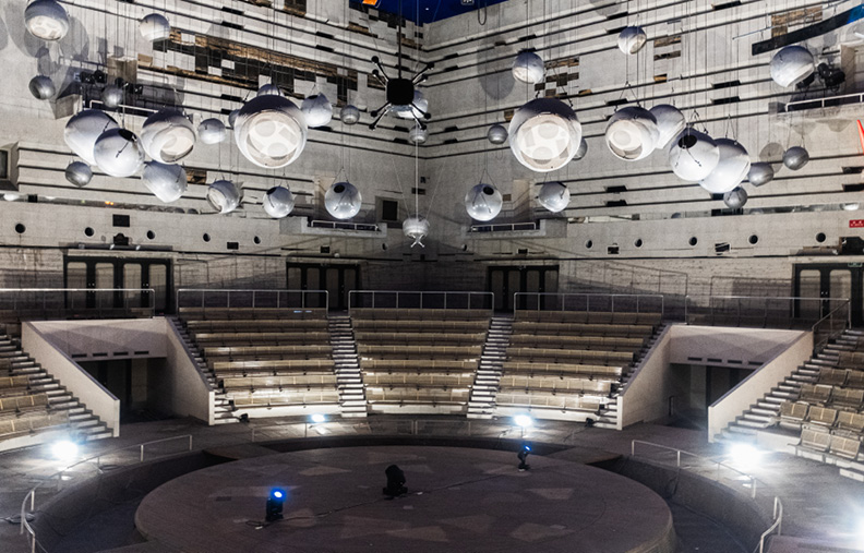 An empty, circular performance stage surrounded by tiered seating in an expansive, industrial-style venue. Large, spherical lights hang from the high ceiling, casting a soft glow over the modern, concrete-and-metal décor.
