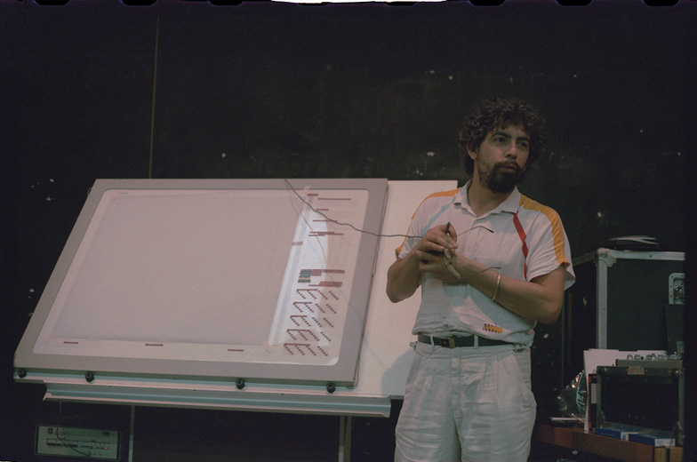 Alain Despres with curly hair and a beard stands in front of the large drawing board of the UPIC. He is wearing a white shirt with colored stripes and white pants. His hands are clasped together, and he is looking slightly to the right. Behind him is a blackboard and some flight cases.