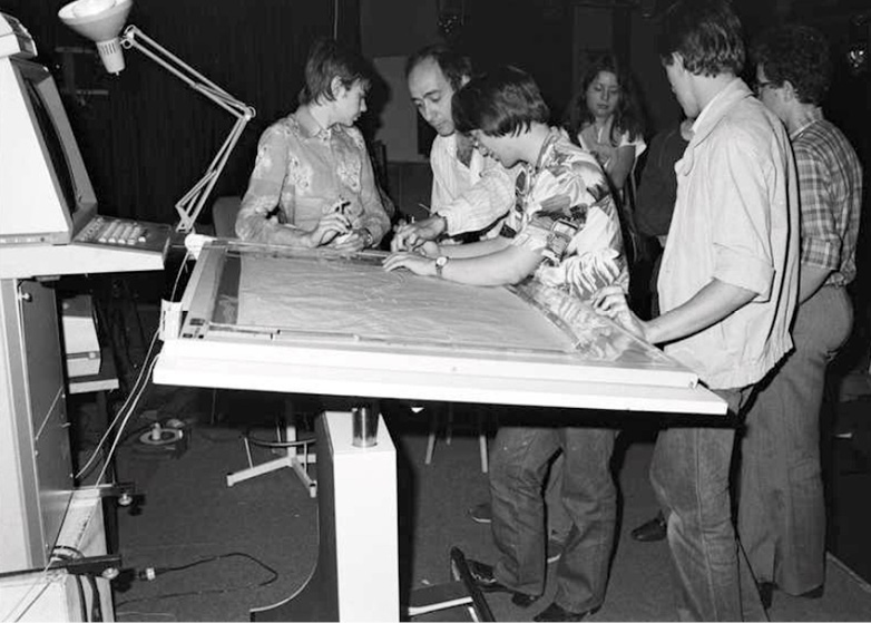 A group of people are gathered around the UPIC drawing table, examining a large UPIC score. The scene is a studio with a lamp, computer, and other office equipment visible. The image is black and white, depicting an older setting.