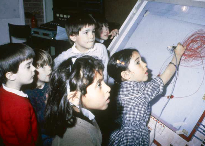 A group of young children gather around the large, angled drawing board of the UPIC. One girl is using a stylus to draw on the surface, illustrating with red lines. The children appear focused and engaged, watching intently as she draws.